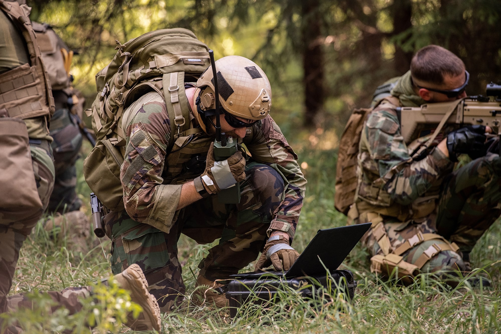 Busy soldier with backpack using military laptop while passing information through radio device in forest