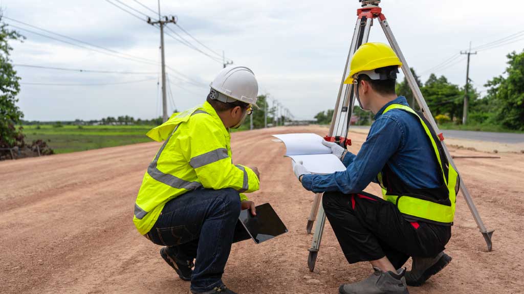 Asian engineer holding tablet to inspect work with foreman using surveying equipment to check pin against road plan.